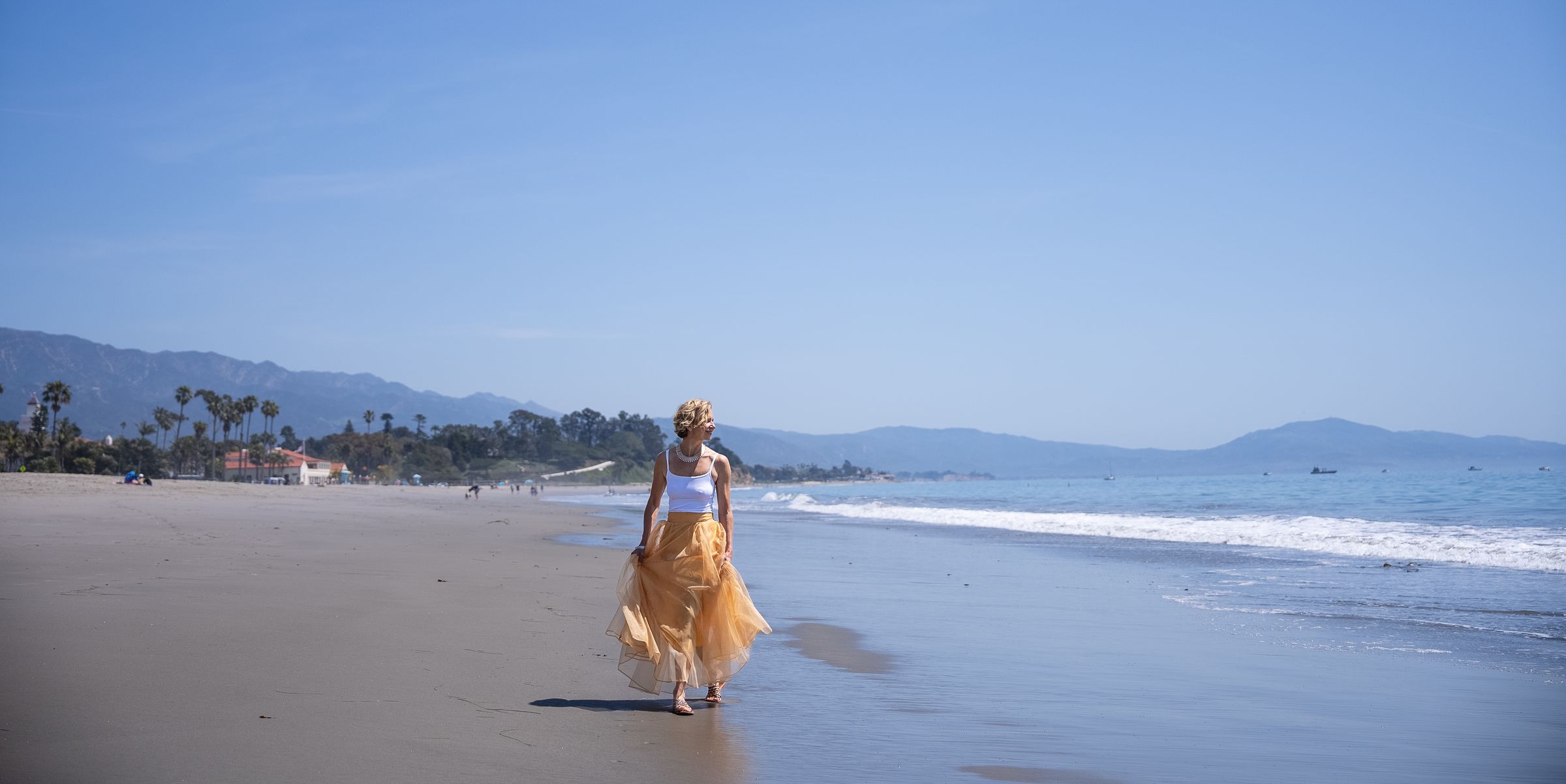 Michelle walking barefoot along the shoreline in a flowing gold skirt, looking back over her shoulder